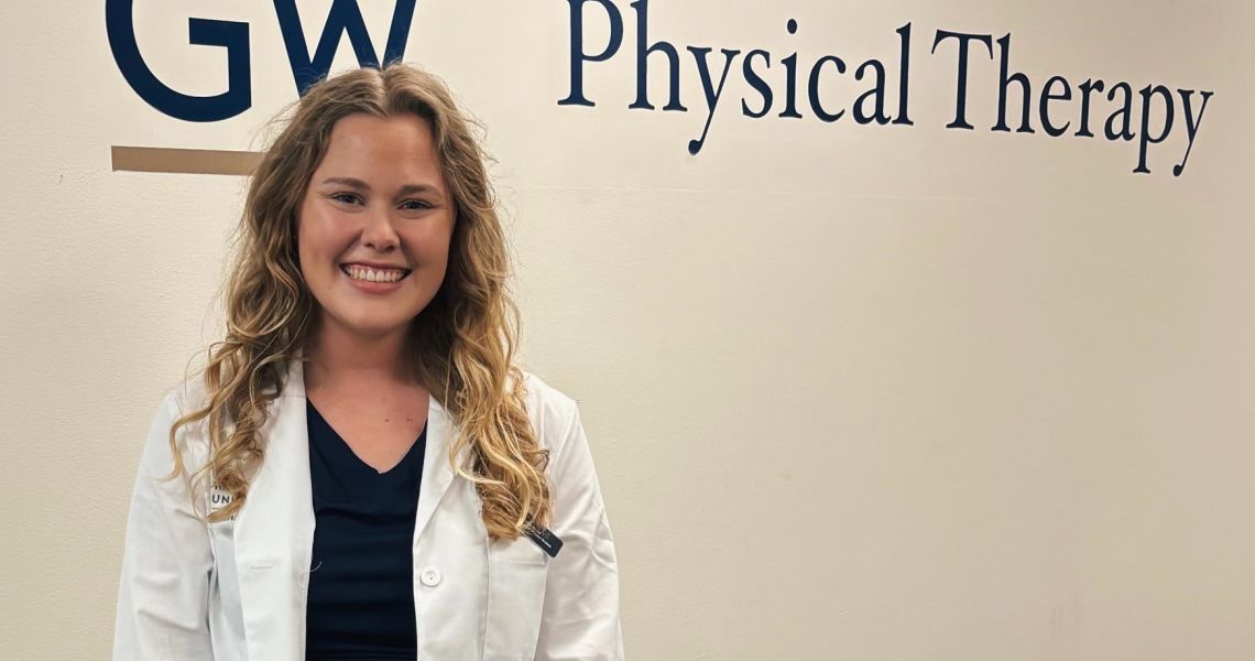 Blonde haired woman in whitecoat standing in front of GW PT sign