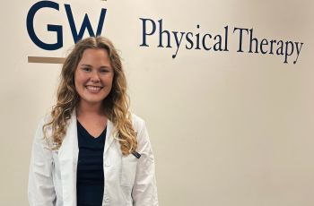 Blonde haired woman in whitecoat standing in front of GW PT sign